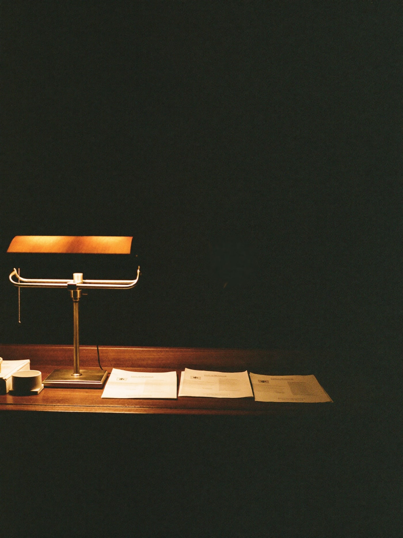 A hotel front desk before dawn — one brass banker's lamp lit low over printed guest lists spread across the walnut counter, the rest of the room still dark.