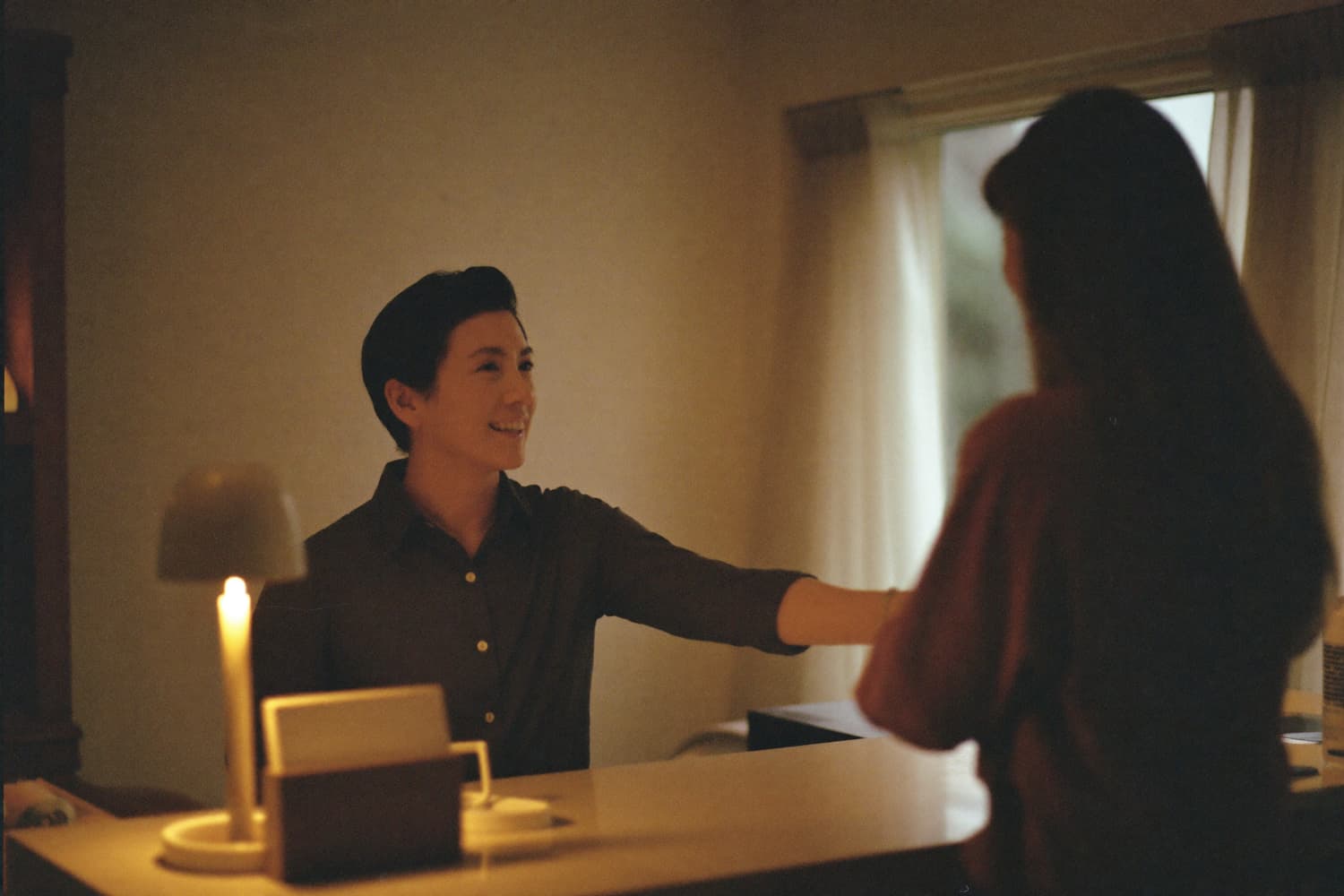 A host welcoming a guest at the desk, unhurried and fully present, no paper in sight.